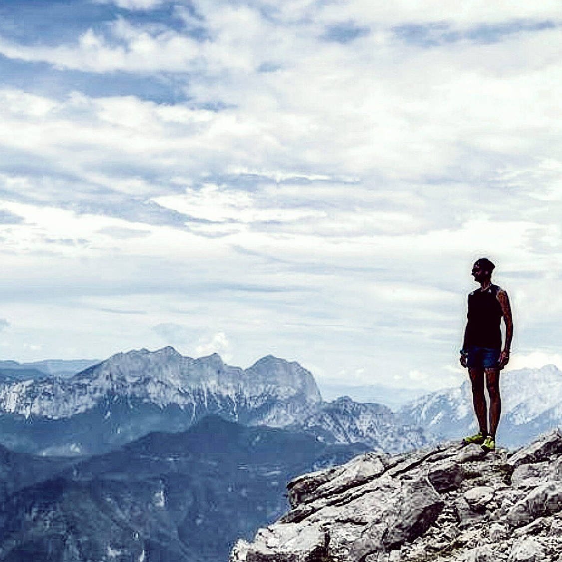 Eine Person steht auf einer Felsklippe und genießt eine innere Reise, während sie unter einem wolkigen Himmel auf ferne Berge blickt. Die Szene erinnert an Gewahrsein und verbindet Abenteuer und Einsamkeit in dieser hochgelegenen, natürlichen Umgebung.