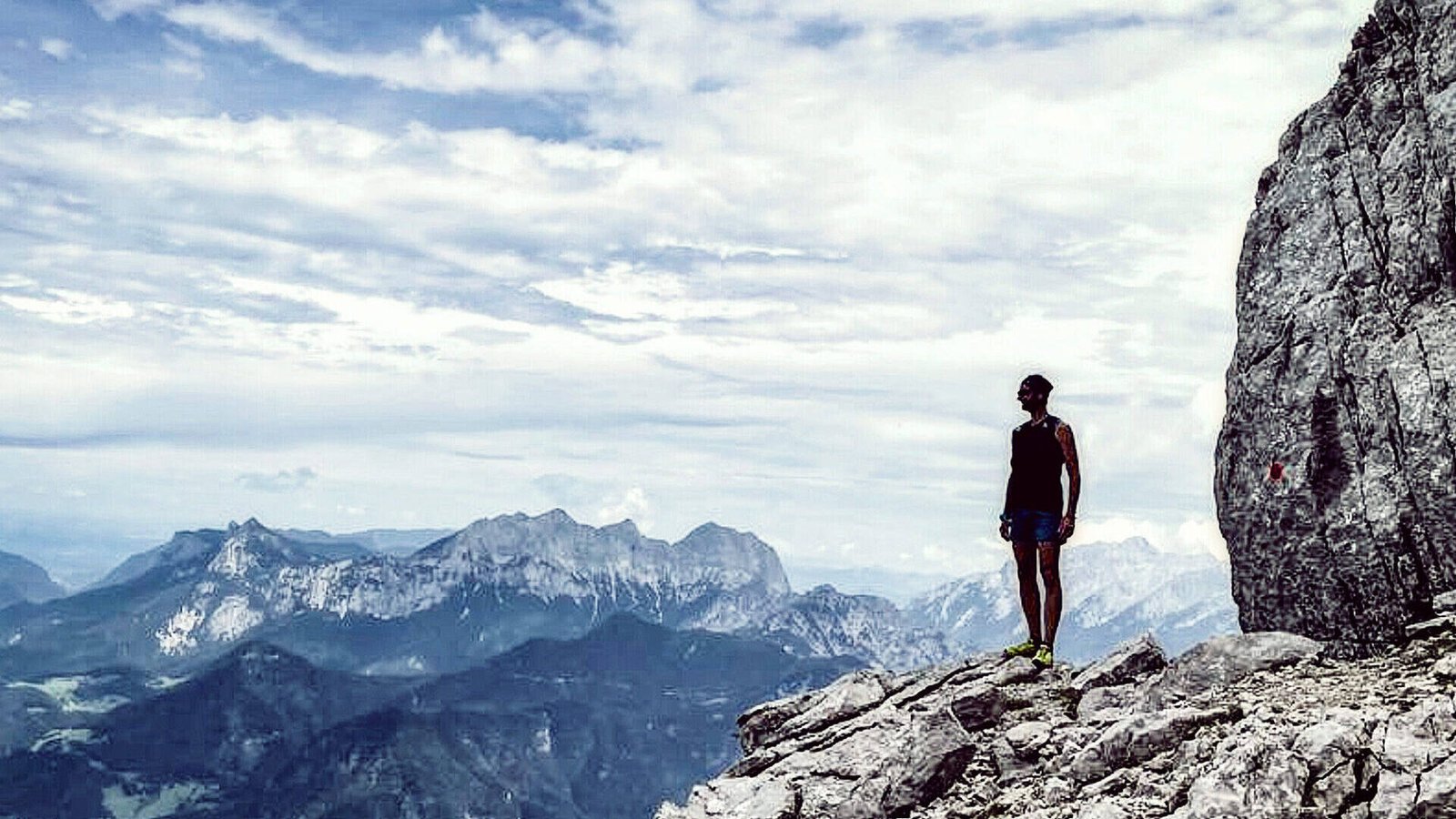 Eine Person steht auf einer Felsklippe und genießt eine innere Reise, während sie unter einem wolkigen Himmel auf ferne Berge blickt. Die Szene erinnert an Gewahrsein und verbindet Abenteuer und Einsamkeit in dieser hochgelegenen, natürlichen Umgebung.