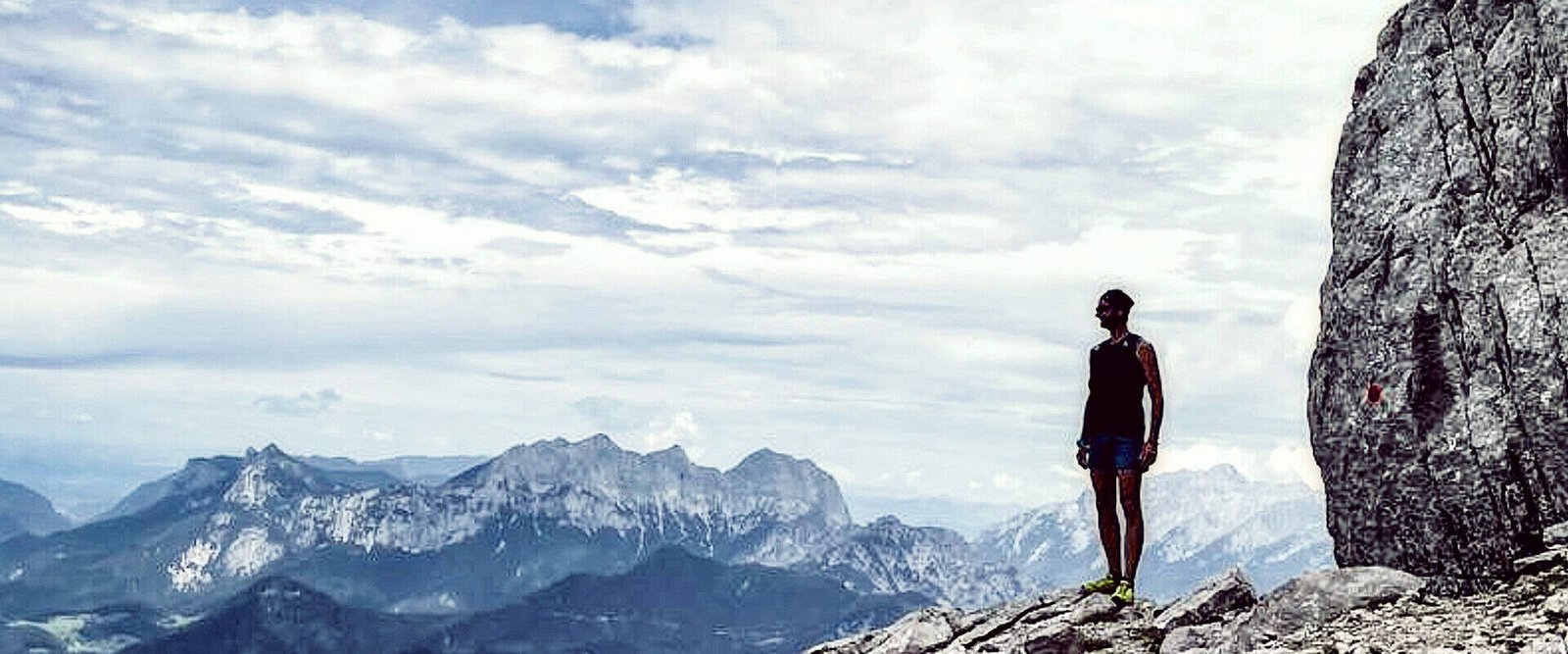 Eine Person steht auf einer Felsklippe und genießt eine innere Reise, während sie unter einem wolkigen Himmel auf ferne Berge blickt. Die Szene erinnert an Gewahrsein und verbindet Abenteuer und Einsamkeit in dieser hochgelegenen, natürlichen Umgebung.