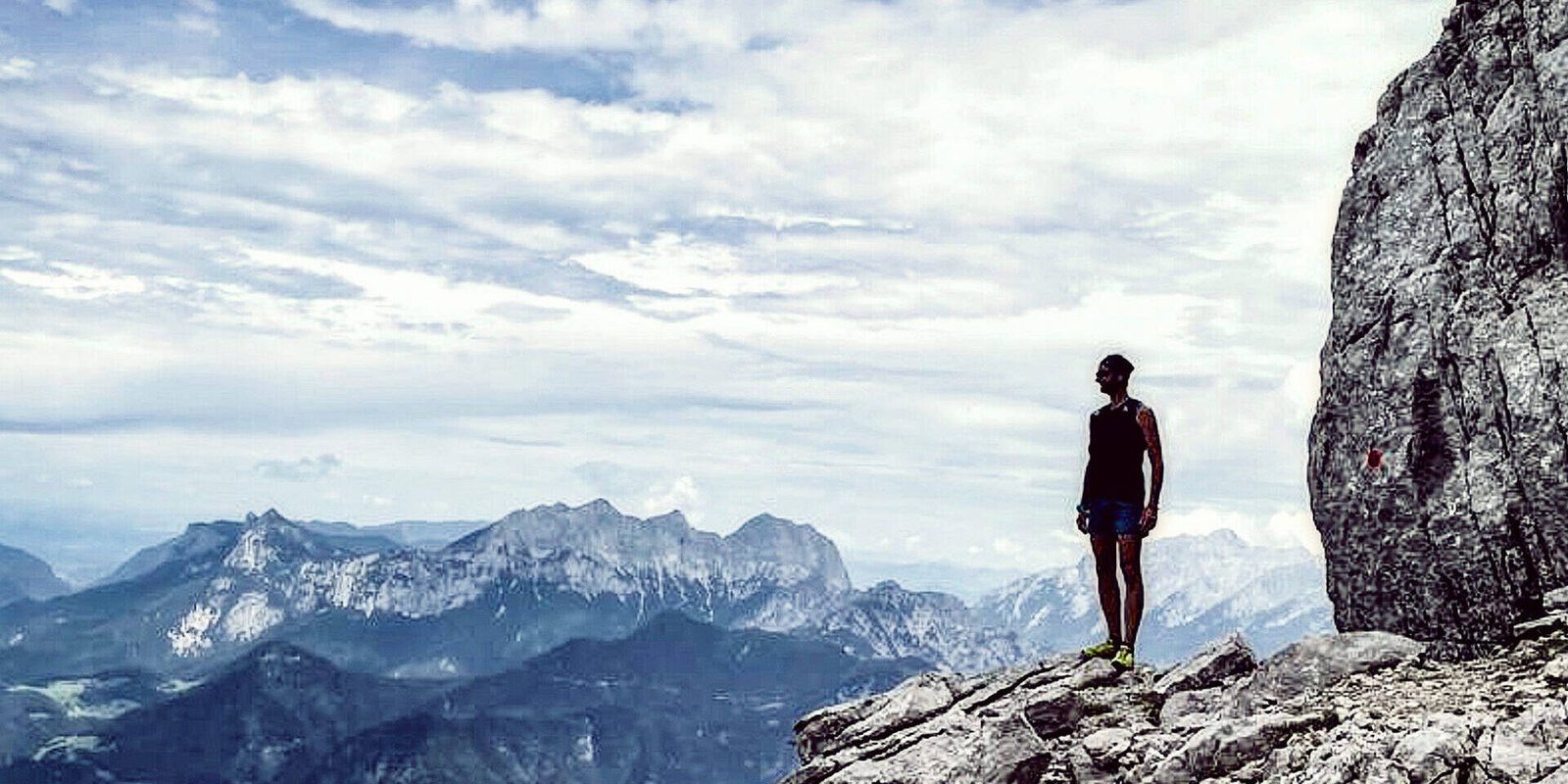 Eine Person steht auf einer Felsklippe und genießt eine innere Reise, während sie unter einem wolkigen Himmel auf ferne Berge blickt. Die Szene erinnert an Gewahrsein und verbindet Abenteuer und Einsamkeit in dieser hochgelegenen, natürlichen Umgebung.