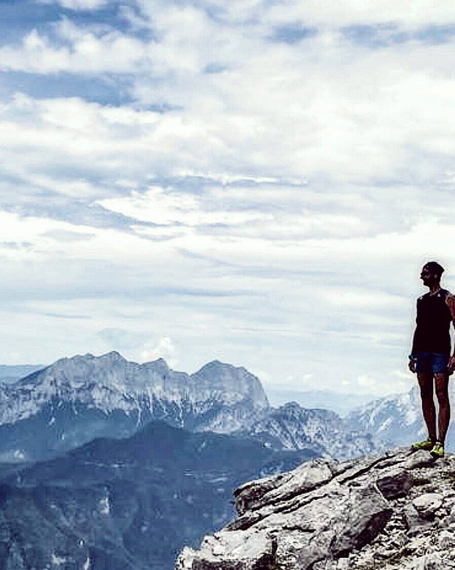 Eine Person steht auf einer Felsklippe und genießt eine innere Reise, während sie unter einem wolkigen Himmel auf ferne Berge blickt. Die Szene erinnert an Gewahrsein und verbindet Abenteuer und Einsamkeit in dieser hochgelegenen, natürlichen Umgebung.