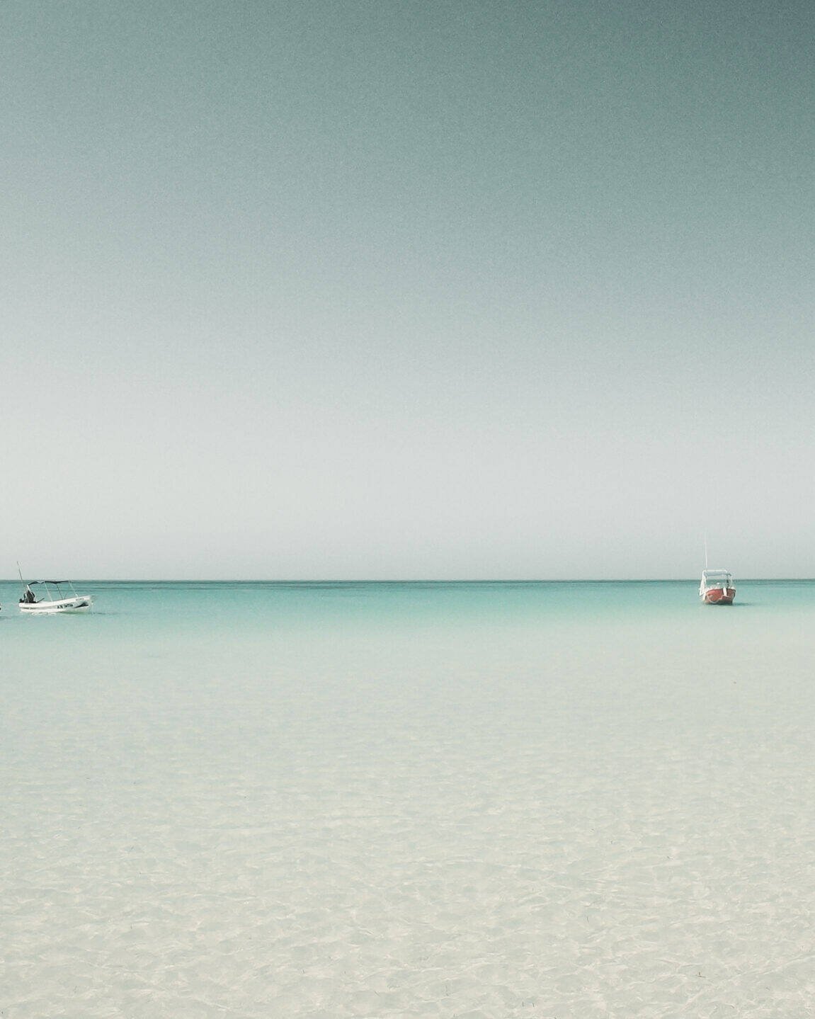 Drei kleine Boote schwimmen auf ruhigem, klarem türkisfarbenem Wasser unter einem blassblauen Himmel und spiegeln eine innere Reise wider. Der Horizont ist sichtbar und das Meer ist ruhig, was eine heitere und friedliche Szene schafft, in der man über die Essenz des Lebens nachdenken kann.