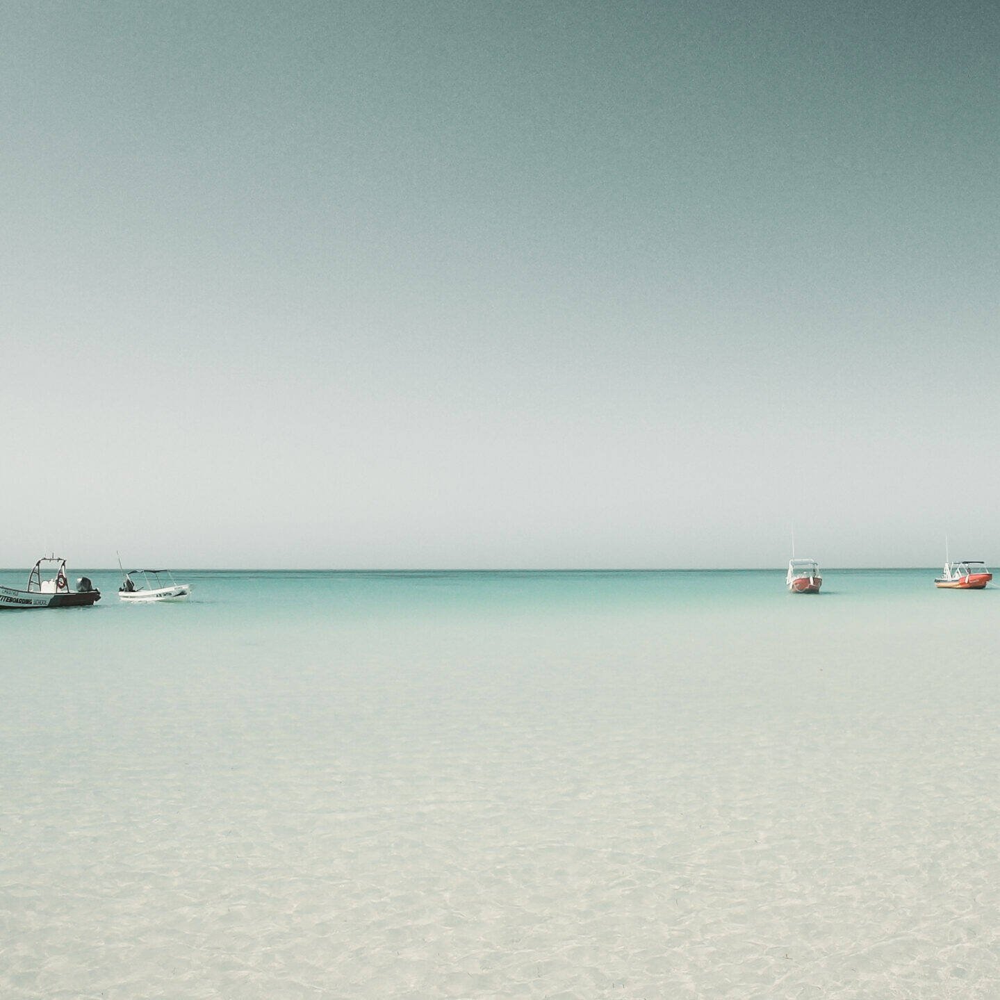 Drei kleine Boote schwimmen auf ruhigem, klarem türkisfarbenem Wasser unter einem blassblauen Himmel und spiegeln eine innere Reise wider. Der Horizont ist sichtbar und das Meer ist ruhig, was eine heitere und friedliche Szene schafft, in der man über die Essenz des Lebens nachdenken kann.