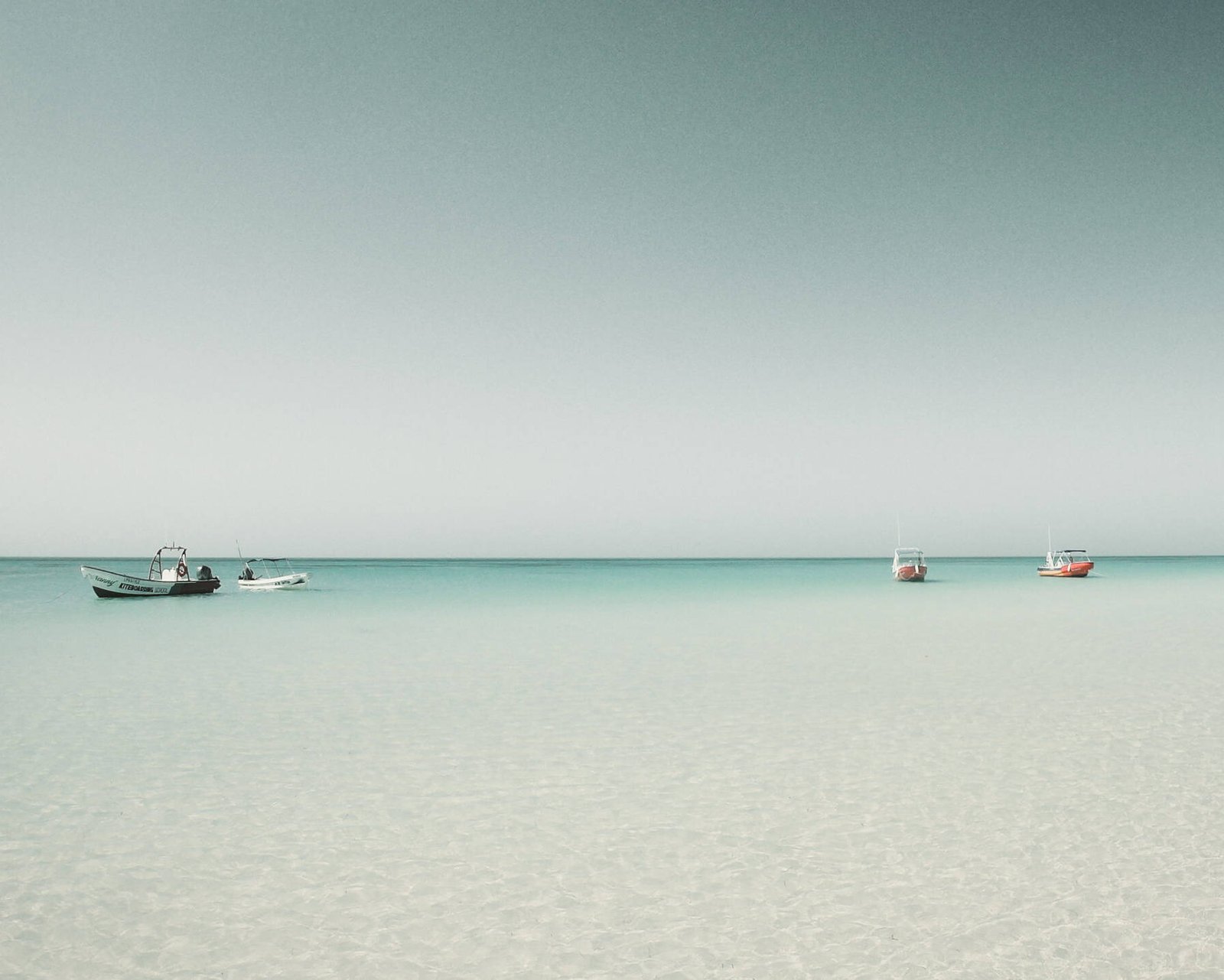 Drei kleine Boote schwimmen auf ruhigem, klarem türkisfarbenem Wasser unter einem blassblauen Himmel und spiegeln eine innere Reise wider. Der Horizont ist sichtbar und das Meer ist ruhig, was eine heitere und friedliche Szene schafft, in der man über die Essenz des Lebens nachdenken kann.