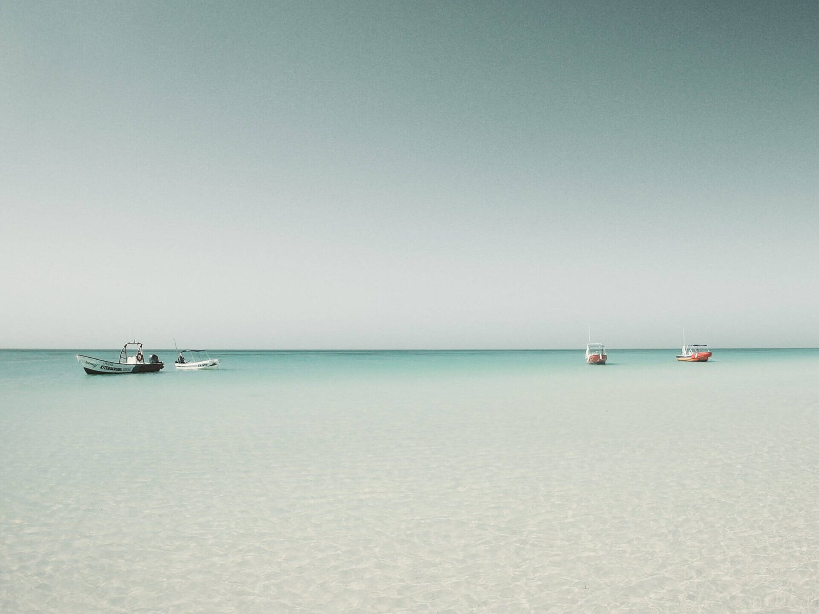 Drei kleine Boote schwimmen auf ruhigem, klarem türkisfarbenem Wasser unter einem blassblauen Himmel und spiegeln eine innere Reise wider. Der Horizont ist sichtbar und das Meer ist ruhig, was eine heitere und friedliche Szene schafft, in der man über die Essenz des Lebens nachdenken kann.