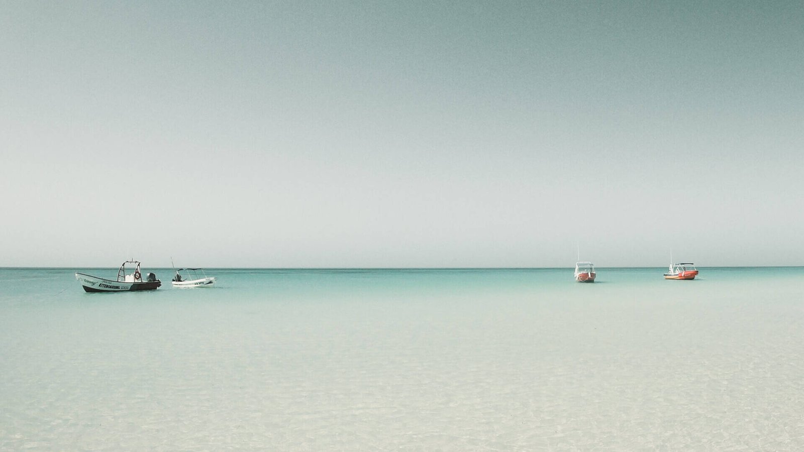 Drei kleine Boote schwimmen auf ruhigem, klarem türkisfarbenem Wasser unter einem blassblauen Himmel und spiegeln eine innere Reise wider. Der Horizont ist sichtbar und das Meer ist ruhig, was eine heitere und friedliche Szene schafft, in der man über die Essenz des Lebens nachdenken kann.