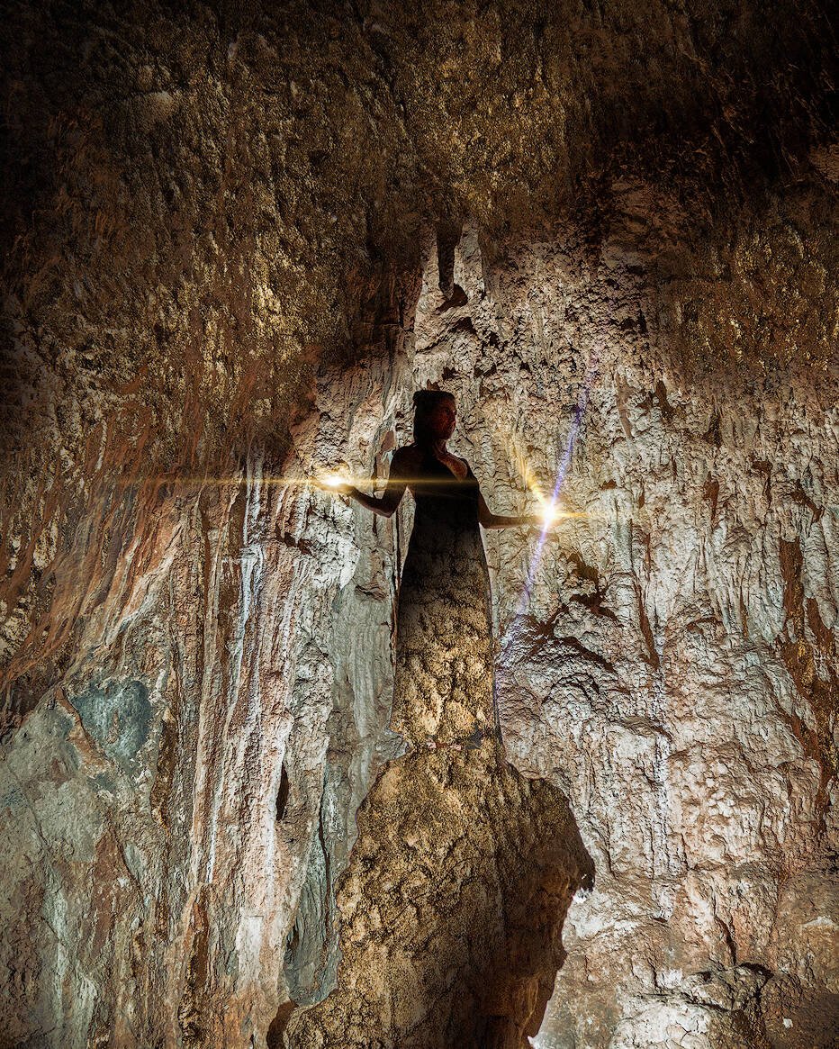 Eine Frau steht in einer schwach beleuchteten Höhle, die von einer einzigen hellen Lichtquelle erhellt wird. Die rauen, strukturierten Wände und die Decke der Höhle umgeben die Figur. Die Szene hat eine ätherische und geheimnisvolle Atmosphäre, als ob sie von Zauber und alter Magie berührt wäre.