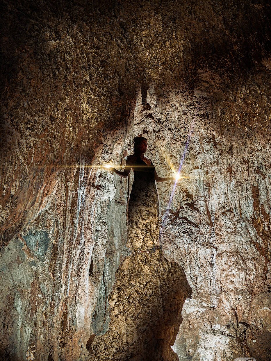 Eine Frau steht in einer schwach beleuchteten Höhle, die von einer einzigen hellen Lichtquelle erhellt wird. Die rauen, strukturierten Wände und die Decke der Höhle umgeben die Figur. Die Szene hat eine ätherische und geheimnisvolle Atmosphäre, als ob sie von Zauber und alter Magie berührt wäre.