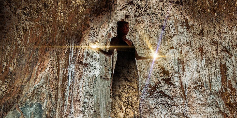 Eine Frau steht in einer schwach beleuchteten Höhle, die von einer einzigen hellen Lichtquelle erhellt wird. Die rauen, strukturierten Wände und die Decke der Höhle umgeben die Figur. Die Szene hat eine ätherische und geheimnisvolle Atmosphäre, als ob sie von Zauber und alter Magie berührt wäre.