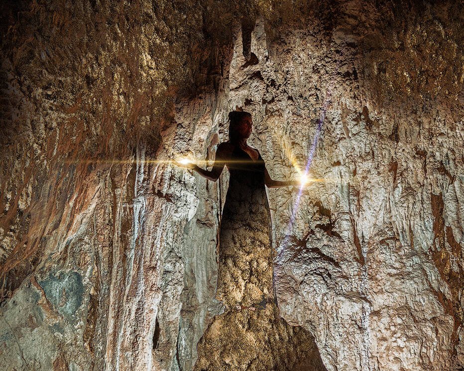 Eine Frau steht in einer schwach beleuchteten Höhle, die von einer einzigen hellen Lichtquelle erhellt wird. Die rauen, strukturierten Wände und die Decke der Höhle umgeben die Figur. Die Szene hat eine ätherische und geheimnisvolle Atmosphäre, als ob sie von Zauber und alter Magie berührt wäre.