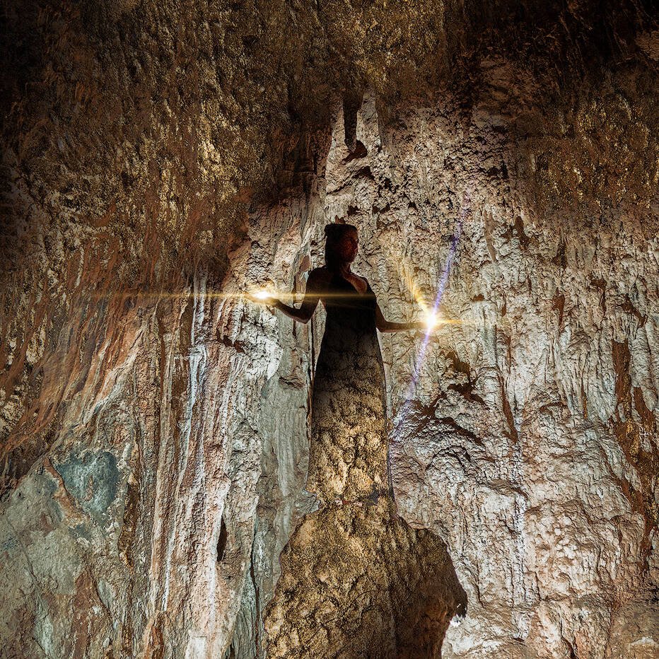 Eine Frau steht in einer schwach beleuchteten Höhle, die von einer einzigen hellen Lichtquelle erhellt wird. Die rauen, strukturierten Wände und die Decke der Höhle umgeben die Figur. Die Szene hat eine ätherische und geheimnisvolle Atmosphäre, als ob sie von Zauber und alter Magie berührt wäre.