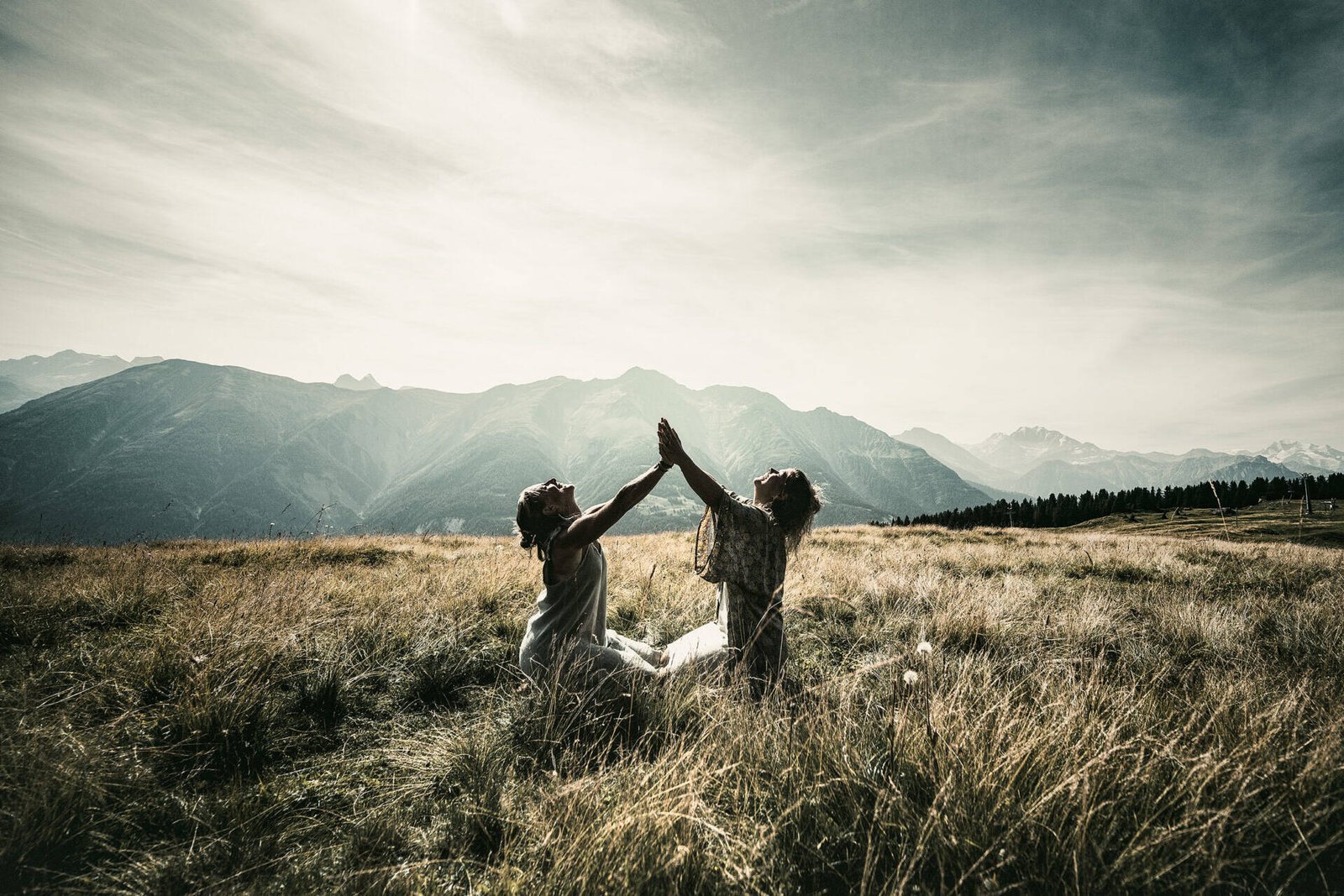 inquiry - zen - anoor_ Zwei Menschen knien auf einer Wiese, einander zugewandt, mit erhobenen Armen und sich berührenden Händen, vor dem Hintergrund der fernen Berge unter einem bewölkten Himmel.