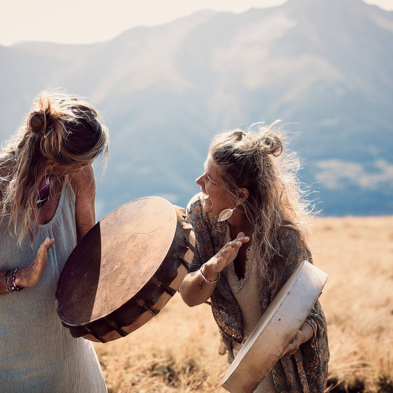 inquiry - zen - anoor_ Zwei Frauen stehen in einer sonnenbeschienenen Wiese mit Bergen im Hintergrund, spielen auf Handtrommeln und lächeln sich an, gekleidet in legere, böhmische Kleidung.