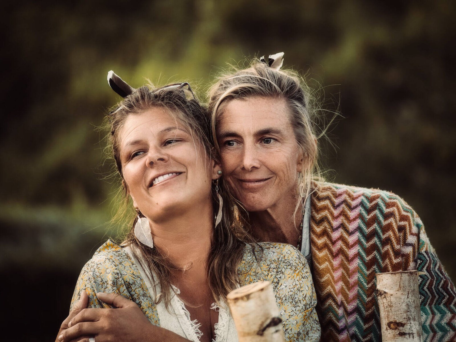 inquiry - zen - anoor_ Zwei Frauen, möglicherweise Mutter und Tochter, stehen im Freien dicht beieinander und lächeln sich freundlich an. Sie haben beide helles Haar und tragen gemusterte Schals. Der Hintergrund ist unscharf und grün, was auf eine natürliche Umgebung hindeutet.