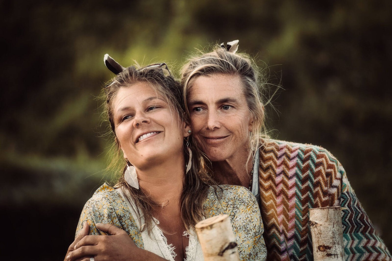inquiry - zen - anoor_ Zwei Frauen, möglicherweise Mutter und Tochter, stehen im Freien dicht beieinander und lächeln sich freundlich an. Sie haben beide helles Haar und tragen gemusterte Schals. Der Hintergrund ist unscharf und grün, was auf eine natürliche Umgebung hindeutet.