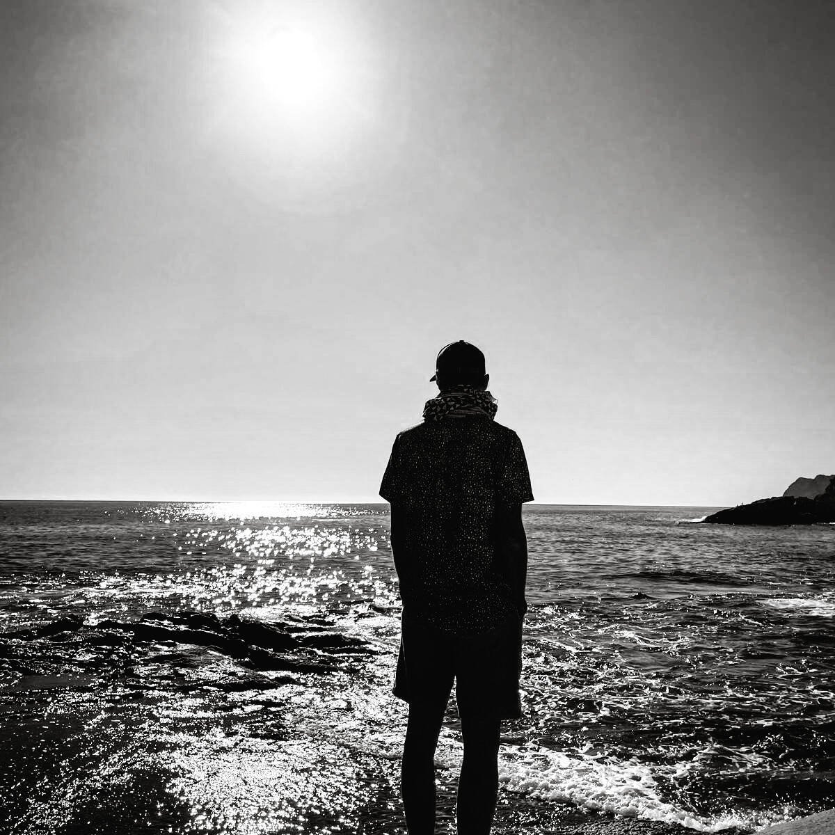 inquiry - zen - anoor_ Eine Person steht an einer felsigen Küste und blickt auf das glitzernde Meer unter einer hellen Sonne am klaren Himmel. Die Szene ist in Schwarz-Weiß gehalten und vermittelt eine friedliche, kontemplative Stimmung.