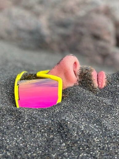 inquiry - zen - anoor_ Das Gesicht einer Person, die teilweise im Sand vergraben ist und eine große gelbe Sonnenbrille mit rosafarbenen, reflektierenden Gläsern trägt. Nur die Nase, die Lippen, ein Teil des Schnurrbarts und die Brille sind über dem Sand sichtbar.