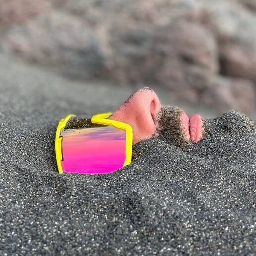 inquiry - zen - anoor_ Das Gesicht einer Person, die teilweise im Sand vergraben ist und eine große gelbe Sonnenbrille mit rosafarbenen, reflektierenden Gläsern trägt. Nur die Nase, die Lippen, ein Teil des Schnurrbarts und die Brille sind über dem Sand sichtbar.
