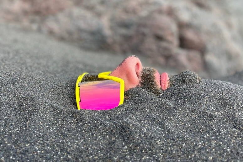 inquiry - zen - anoor_ Das Gesicht einer Person, die teilweise im Sand vergraben ist und eine große gelbe Sonnenbrille mit rosafarbenen, reflektierenden Gläsern trägt. Nur die Nase, die Lippen, ein Teil des Schnurrbarts und die Brille sind über dem Sand sichtbar.