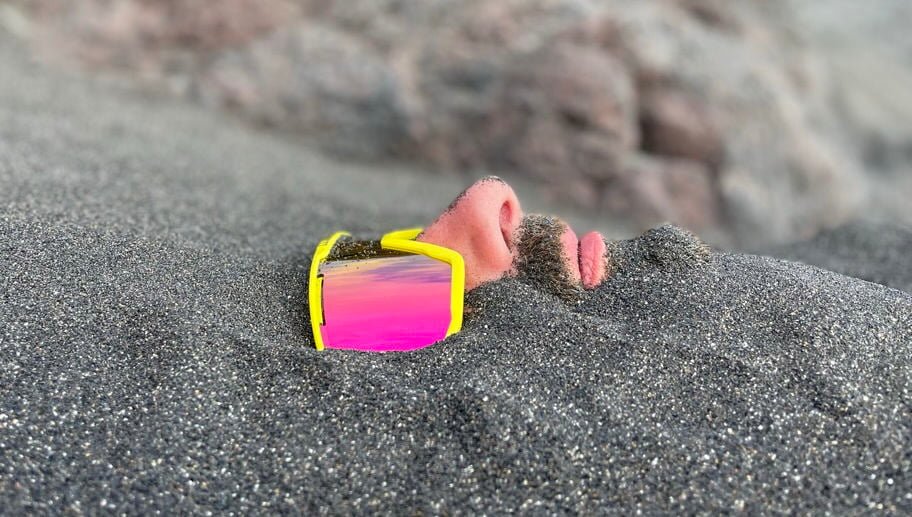inquiry - zen - anoor_ Das Gesicht einer Person, die teilweise im Sand vergraben ist und eine große gelbe Sonnenbrille mit rosafarbenen, reflektierenden Gläsern trägt. Nur die Nase, die Lippen, ein Teil des Schnurrbarts und die Brille sind über dem Sand sichtbar.